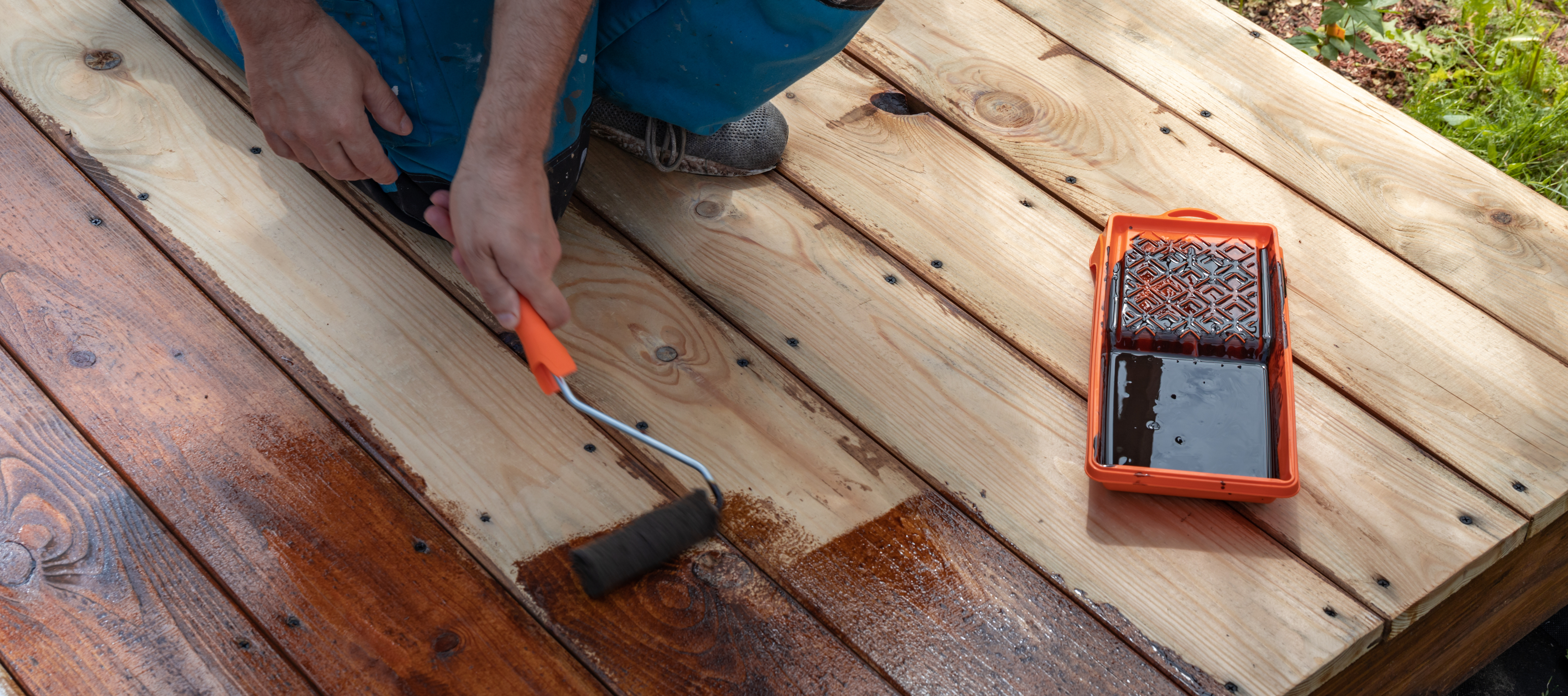 Worker applying wood stain with brush to deck boards next to orange paint tray